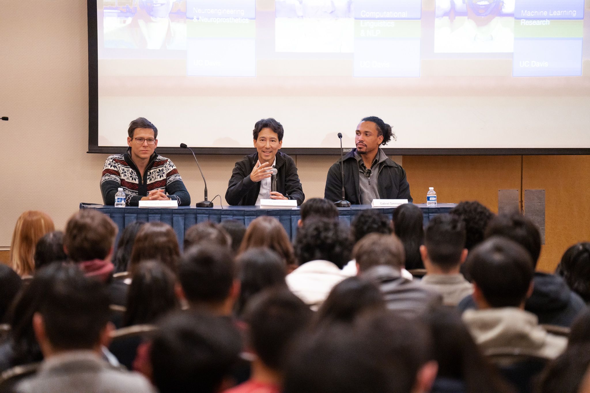 Three men sitting at a table with microphones in front of a crowd